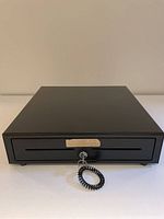 Closed APG cash drawer showing front lock with key and coiled keyring attached.