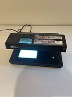 Front left angle showing power cord, viewing window with UV light on, and instructional label.