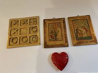 Photo showing brass tic tac toe board with X and O pieces, two wooden framed pictures with vintage artwork, and a red ceramic heart decoration.