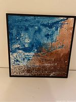 Front view of the square abstract painting with blue, brown, and white colors in black frame.