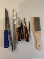 Photo showing the full set of jewelry making tools, including metal file with blue handle, two mandrels, three wooden handled tools, and wire brush