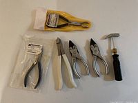 Top down view of six tools laid out, including pliers with white handles, two metal pliers, one hammer with black handle, and one packaged tool with black handles.