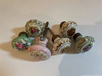 Group of six painted porcelain door knobs with floral and gold accent designs lying on a flat surface. Four knobs have visible floral patterns; two white with gold accents and floral details, one green base with pink roses, one pink base with floral pattern.