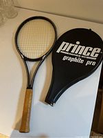 Graphite tennis racket with leather handle and matching cover on white table.