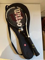 Wilson tennis racquet in black and white with red Wilson logo strings, resting on black Wilson cover with shoulder strap.