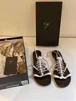 Pair of white fishbone strap women's sandals with brown footbed shown next to original black box with logo and accompanying paperwork inside.