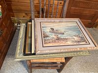 Stack of framed artworks on wooden chair showing the top piece with a crab and shore scene in a large wooden frame.