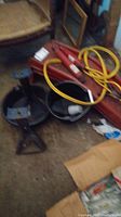 Photo showing a metal jack stand, yellow jumper cables, red Napa shocks, and red car ramps arranged on a garage floor with a black bucket.