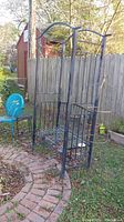 Outdoor photo of the metal garden bench with the attached arched trellis, placed on a brick-paved yard.