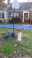 Outdoor setting with concrete bird bath, shepherd's hook, and multiple planter pots arranged around a patch of greenery in front of a house.
