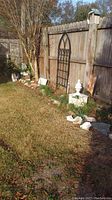 Full view of garden area showing metal garden trellis with gothic arch design, birdhouse mounted on wooden post, white shell ornaments on fence, and a decorative white finial on base