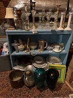 Overview photo showing multiple candlestick holders, plates, bowls, mugs, decorative glass heads, and other items arranged on blue shelving and floor in attic space.