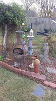 Wide shot of garden area showing chiminea with metal bird decoration, religious statue, plant stands, various pots, and garden ornaments in a backyard setting.