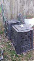 Two black resin composters with vent holes and lids, placed outdoors next to a wooden fence. Wire mesh fencing rolls and stakes are visible behind them.