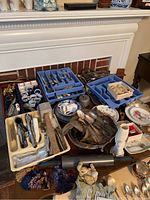 Wide view of assorted silver plated cutlery in trays along with serving items, ceramic plates, napkin rings, and other kitchenware on display on a table in front of a fireplace.