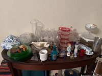 Wide view of assorted glassware and ceramics on wooden table showing clear glass pitcher, blue and white butter dish, green plates, candyst dishes, stemware, figurines, and other small items