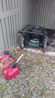 Full view of lawn mower next to two red gas cans on leaf-covered ground outside a shed.