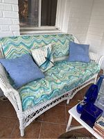 Full view of the white wicker loveseat with blue-green patterned seat cushion, two blue pillows, and one beige decorative pillow with dragonfly and script design. Shows overall condition and setup.