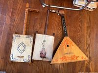 Three string instruments on hardwood floor: two rectangular frame instruments with stretched membranes and one triangular wooden string instrument with painted orange decoration.