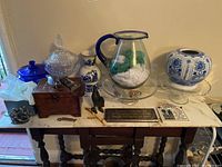 Overview of items on marble-top wooden table including glass pitcher, blue and white vases, glass canisters, wooden box, animal figurines, plaques.