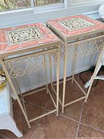 Two cream-colored metal frame plant stands with square terracotta and cream pottery tops showing wear and decorative details.