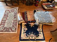 Image showing multiple rugs and rug runners on a wooden floor along with a folded flag. Rugs include various sizes and patterns, some rolled or folded and one wrapped in plastic.