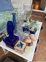 Top view of white table holding multiple blue and clear glass vases, bowls, decorative tiles and sea shells.