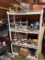 Four-tier plastic shelf with kitchenware items including Blue Danube plates, bowls, goblets, cups, and ceramic eggs arranged on shelves in an attic area.