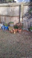 Outdoor photo of terracotta pots, blue ceramic pots, watering cans, and bottles arranged on grass near a wooden fence.