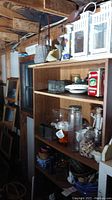 Full shelf showing white wooden lanterns, clear glass bottles, jars with lids, bowls, and metal decorative items on the wooden shelving unit in a garage.