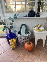Full view of lot showing blue glass vase with faux leaves, translucent glass vase with faux lemons, ceramic pot with detailed design, ceramic orange pumpkin, ceramic basket with shells on white stool, and small blue vase on window ledge.