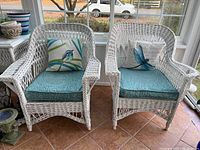 Two white wicker chairs side by side with blue patterned cushions and decorative pillows.