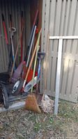 Wide view of yard tools leaning inside a shed and exterior area including a broom, mop, rakes, shovels, wheelbarrow wheel, crowbar, and splitter tool
