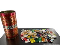 Decorative tall tin container without lid with red background and gold lettering containing matchbooks