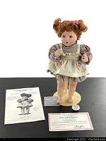 Full shot of Betsy collector doll standing on display stand with accompanying papers and certificate.