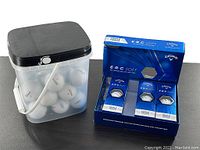 Photo showing bucket of reclaimed golf balls with black lid and white handle, alongside three unopened 3-packs of Callaway E.R.C Soft golf balls in blue packaging.