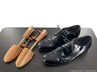 Photo showing both black patent leather dress shoes and wooden shoe stretchers side by side on a dark surface.