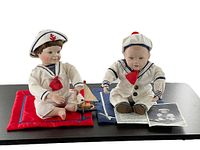 Pair of Ashton Drake sailor dolls seated on cushion, one holding photo and certificate, other sitting beside wooden sailboat toy.