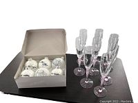 Set of eight champagne flutes with pink frosted stems arranged beside an open box containing six white glass wedding ornaments decorated with black wedding-themed designs and text.