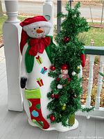 Side angle showing fabric snowman standing next to decorated artificial Christmas tree on a porch.