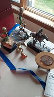 Overall view of the lot showing various bronze and brass figurines including Pony Express rider, child with birds, brass birds, kitten, and red stone carving.