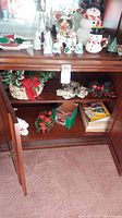 Wide view of wooden cabinet displaying Christmas items such as baskets with faux floral, snowman figurines, wreath, and boxes with decorations inside shelves.