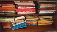 Photo showing stacked hard and softcover cookbooks on shelves and floor, displaying various titles related to cooking, diet, herbs, and recipes.