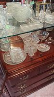 Multiple cut and pressed glass bowls, plates, cups, and trays arranged on a wooden dresser top, showing various patterns and shapes mostly in clear glass.