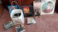 Wide shot showing multiple boxed sets and CDs arranged on carpet including Elvis Presley and others, some inside a white paper bag.