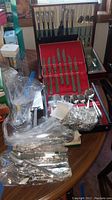 Various stainless steel flatware pieces and sets arranged on table, some in boxes with red lining, others wrapped in plastic
