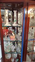 Display cabinet containing a variety of clear glass and crystal candle holders, as well as tall white ceramic candlesticks with green designs on a glass shelf.