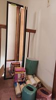 Tall rectangular wall mirror leaning against wall with several rolled bathtub mats, two boxed items, and a teal waste can on the floor nearby.