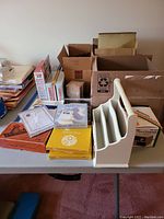 Photo showing office supply items including glue gun in packaging, address books, desk organizer, boxes with pencil sharpener, pen sets, and snap hook.