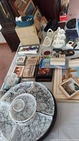Photo showing various items including white ceramic tulip-shaped holders, several boxed Fisher Space Pens, tins with scenic prints, placemats, and a serving tray with a marbled black and white pattern with compartments.